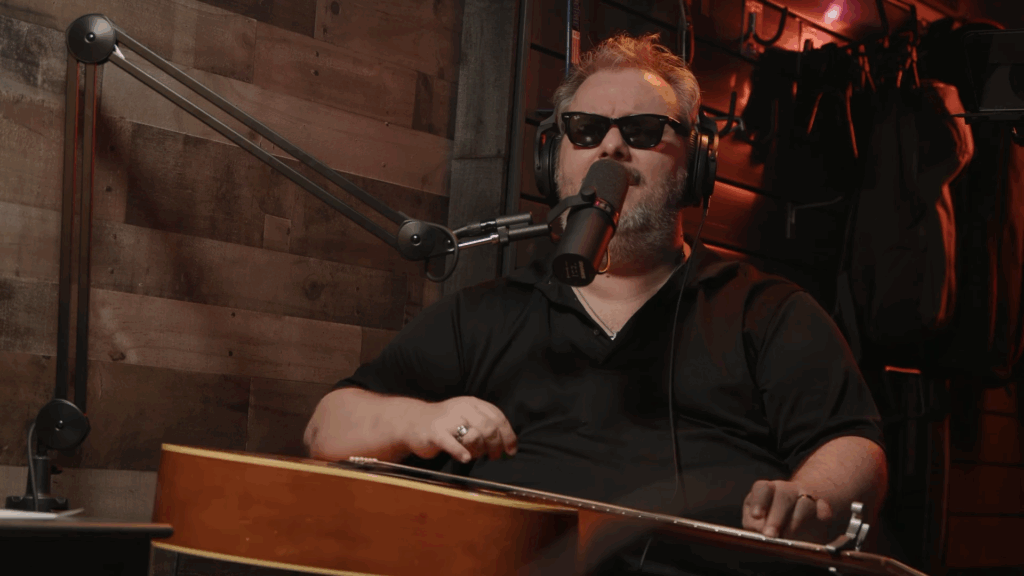 Randy Gilkey sings into a microphone while playing a guitar that lays flat on his lap.